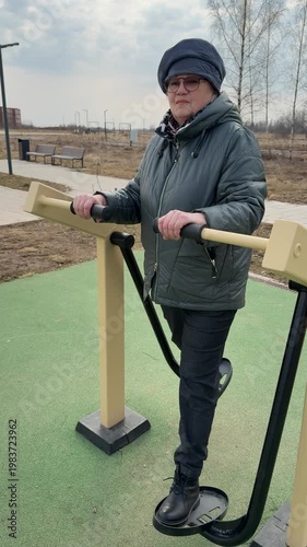 Woman exercising on outdoor leg trainer on playground turf in park during spring day senior performs steady side steps holding.