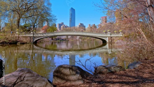 4K static shot of the iconic Bow Bridge in Central Park during early spring. The white cast-iron bridge reflects perfectly in the calm lake water, framed by budding trees and the modern skyline
