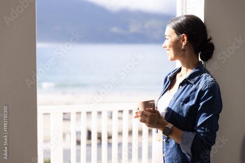 Woman is standing and holding ceramic mug, checking smartwatch on balcony overlooking sandy beach