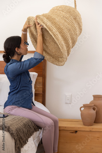 Woven straw pendant shade swinging above neatly made bed with textured throw in bedroom, copy space