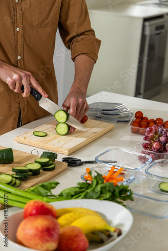 Chef's knife is slicing cucumber on wooden cutting board on white countertop, surrounded by produce