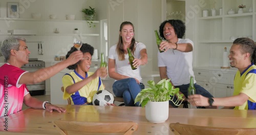 Diverse friends in jerseys toasting bottles after goal holding soccer ball at home kitchen table