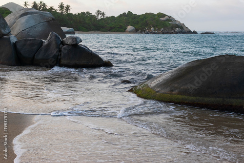 Exotic shoreline landscape with large rounded stones, foamy waves, and rainforest foliage in background