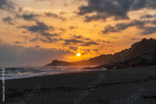 Golden sunset over sandy beach with waves, rocky breakwater, and dramatic cloud formations on horizon