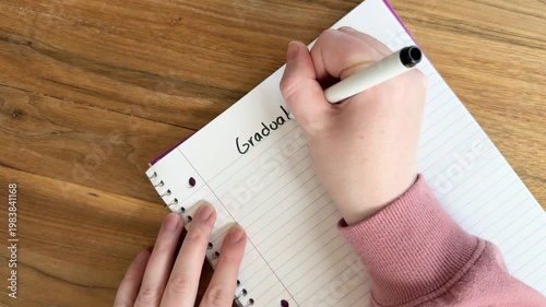 Close up of hands writing “graduation” on lined notebook paper, education milestone and celebration concept, natural light overhead shot on wooden table, 4K video
