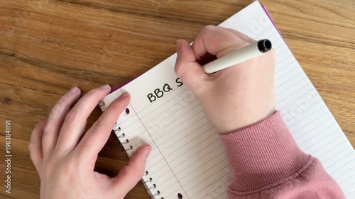 Close up of hands writing “BBQ shopping list” on lined notebook paper, summer grilling and grocery planning concept, natural light overhead shot on wooden table, 4K video