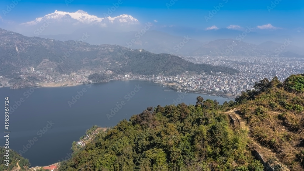 Fototapeta premium The beautiful blue lake is surrounded by mountains. Green forest on the slopes. A city in a valley. Snow-capped peaks of the mountains, azure sky. Pokhara. Lake Phewa. The Himalayas. Annapurna.