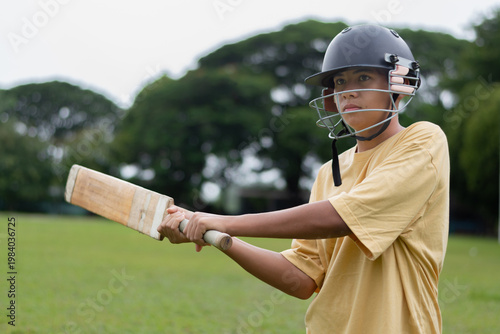 A young boy wearing a helmet and yellow t-shirt playing cricket in a sunny field. He swings the bat to hit cricket ball with focus and energy, enjoying the sport outdoors.