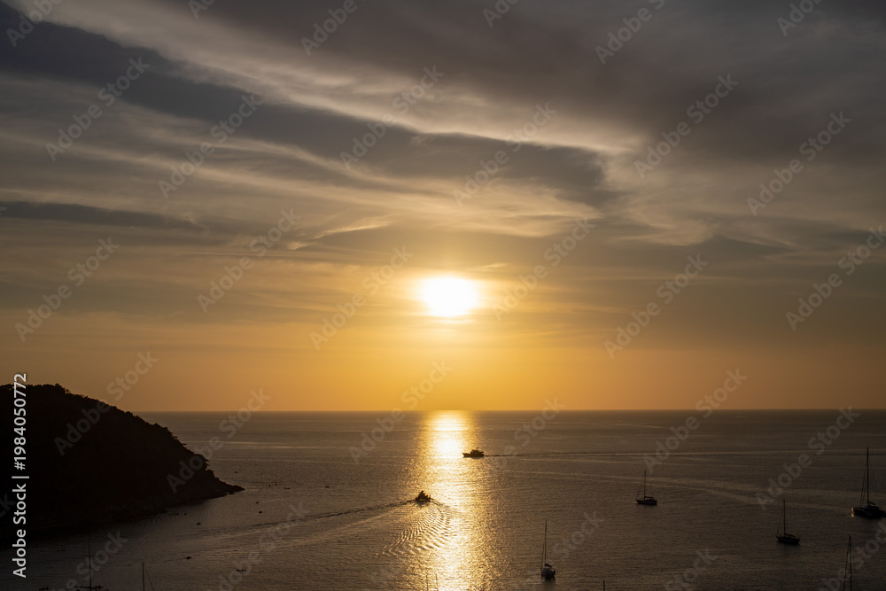 Obraz premium Golden sunset over the Andaman Sea from Windmill Viewpoint in Phuket, with sailboats drifting on calm water and warm reflections stretching across the horizon.