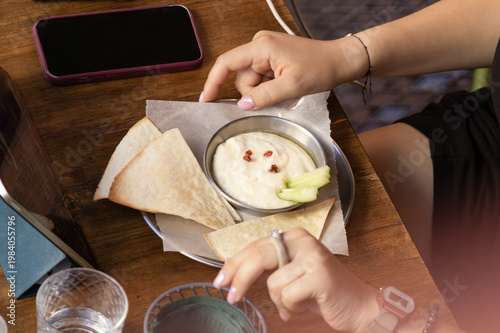 Small plate with flatbread, creamy dip, and cucumber slices sits on wooden table alongside phone, water glass, and hands in relaxed dining setting. Scene conveys informal atmosphere, texture, and ever