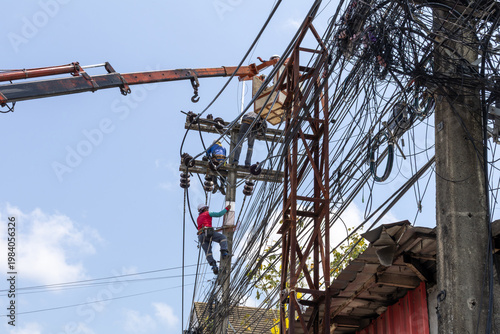 Electricians in safety gear are working on a tangled mass of power lines and a transformer on a utility pole in Thailand. A crane arm is visible, likely assisting with the heavy equipment replacement.