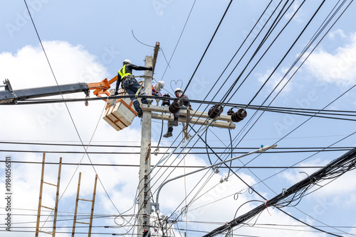 Electricians work on high-voltage power lines and a utility pole in Thailand, using a crane bucket and safety gear while performing maintenance and wiring tasks.