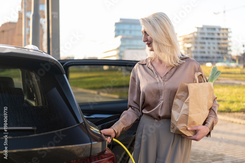 Foto Charging electric vehicle at a public station while loading grocery bags into the trunk