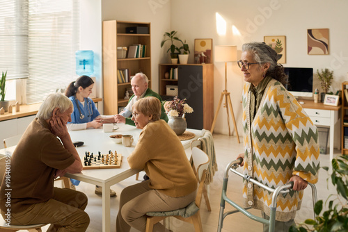 Slika na platnu Senior Caucasian woman with disability using walker while elderly group socializing and playing chess in care center