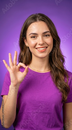 Smiling young woman showing ok hand gesture wearing purple t-shirt on purple background. Approval and positive feedback concept.