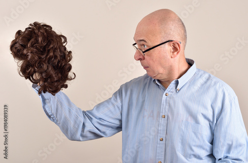 Middle-aged bald man with eyeglasses holding and looking in surprise at a wig. Health or aging problem concept.