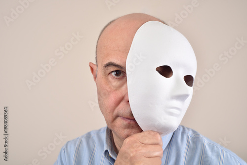 Middle-aged man with sinister smile holding a white mask over half of his face and symbolizing hidden identity.