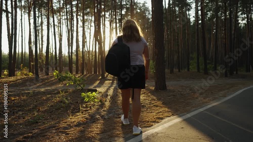 Young woman with backpack walking on forest path at sunset. Back view, sunlight through pine trees, summer nature hike, outdoor activity, solo travel, trekking in woodland.