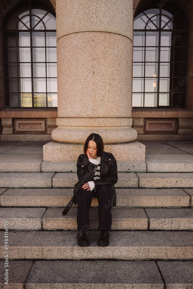 Naklejka premium Stylish woman in leather jacket sitting on stone steps in city