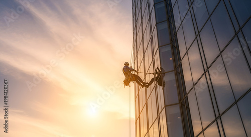 Two people rappelling down a modern glass skyscraper at sunset.