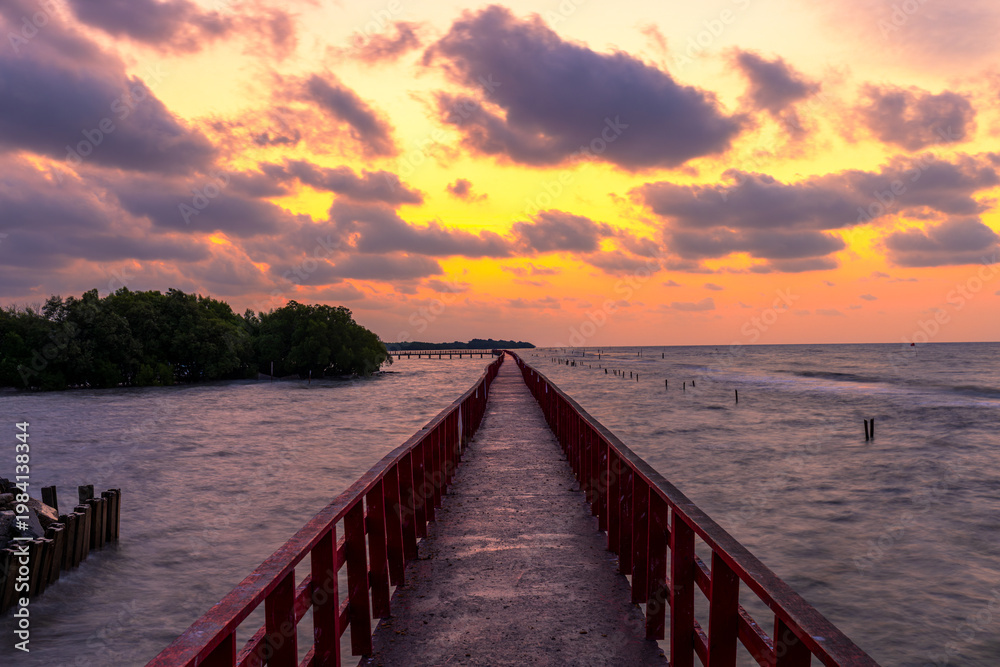 Obraz premium Perspective of Red Wooden Bridge over the Sea at Golden Sunset,A long red wooden bridge stretches into the sea towards the horizon under a dramatic golden sky during sunset. The scene features lush.