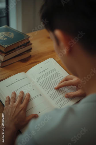 Person reads a book while sitting at a wooden table near a stack of books in a cozy room