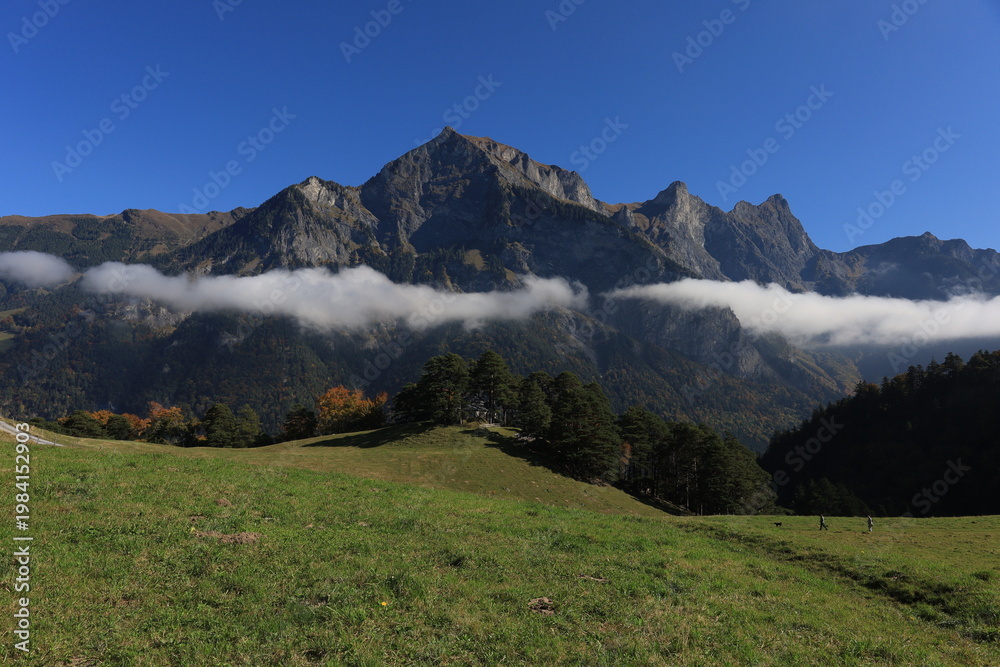 Fototapeta premium Misty peaks rise above green meadows. Clouds drift between rugged mountain ridges. Clear blue sky frames the tranquil scene. Nature's grandeur invites calm reflection