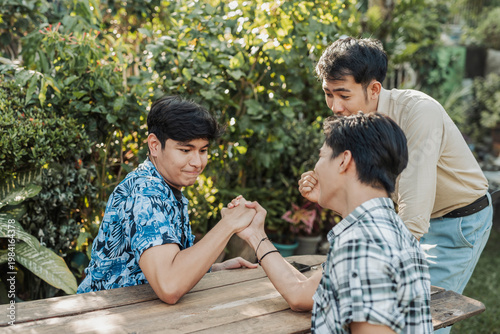 A focused young Asian man mediating between two friends during a playful arm wrestling match while keeping the competition friendly and fun. Outdoor shot.