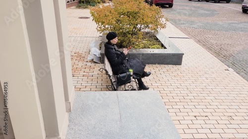 Elegant woman in black beret and coat sitting on a city bench using her smartphone