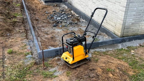 Yellow plate compactor machine positioned on a construction site with freshly leveled soil and a concrete foundation wall in the background