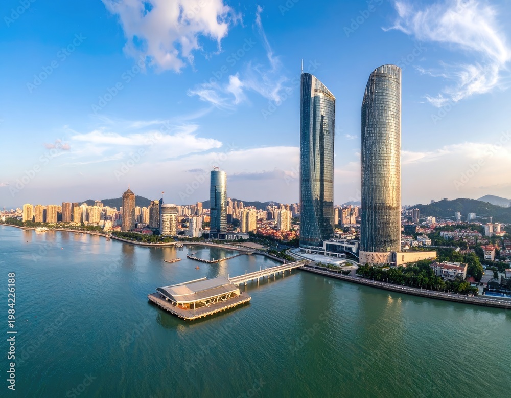 Obraz premium An aerial view showcases the modern skyline of a coastal Chinese city, featuring towering skyscrapers reflected in the calm waters of a bay under a clear blue sky.
