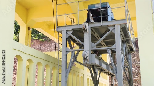 Side view shows industrial gantry and crane mechanism for dam gates. Steel structure stands on rails near yellow concrete wall. Heavy industrial gantry supports the crane mechanism.