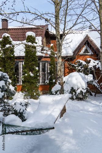 Snowy winter scene with brick house covered in white snow. Tall evergreen shrubs and small decorated tree are surrounded by snow, and snow-laden hammock is visible in foreground.