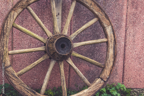 Rustic wooden wagon wheel rests against a textured wall adorned with creeping greenery in a tranquil outdoor setting