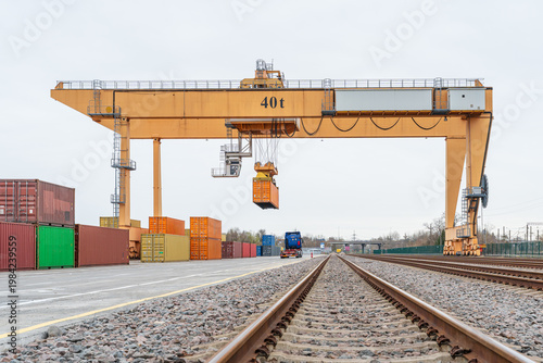Yellow 40t gantry crane lifting an orange container at an intermodal rail terminal, professional heavy-duty logistics equipment for cargo handling and global trade