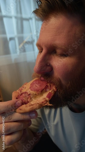 Vertical slowmo closeup shot of bearded young Caucasian man enjoying slice of delicious pizza at home