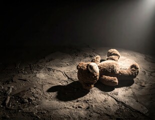 Abandoned teddy bear lying on a dusty ground illuminated by a soft beam of light