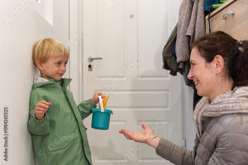 Wallpaper Mural Young boy in green raincoat holding colorful craft supplies smiles at woman in cozy sweater, engaged in playful conversation in a bright indoor setting Torontodigital.ca