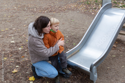 Wallpaper Mural Woman with dark hair, wearing a cozy sweater, embraces a young boy in an orange jacket, both sitting near a playground slide, enjoying a moment of connection in an outdoor setting Torontodigital.ca