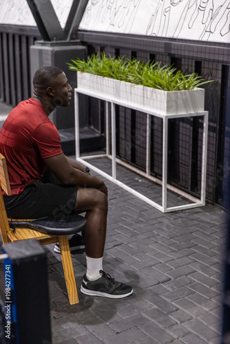Man is resting on wooden bench in modern gym lounge area, with white planter and mural