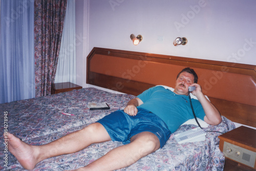 Middle aged man in casual clothes lying on double bed in hotel room talking on corded telephone, analog color film mid 1990s
