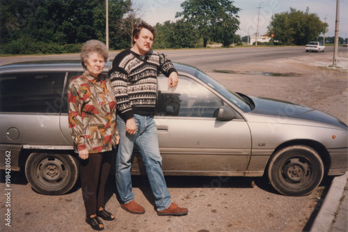 Mother and son standing together next to silver hatchback car on roadside on sunny day, analog color film mid 1990s