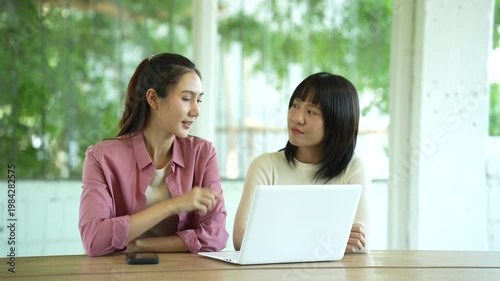 Two young women smiling and collaborating on a laptop in a bright modern workspace, representing teamwork, productivity, and a positive working atmosphere.