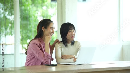 Two young women smiling and collaborating on a laptop in a bright modern workspace, representing teamwork, productivity, and a positive working atmosphere.