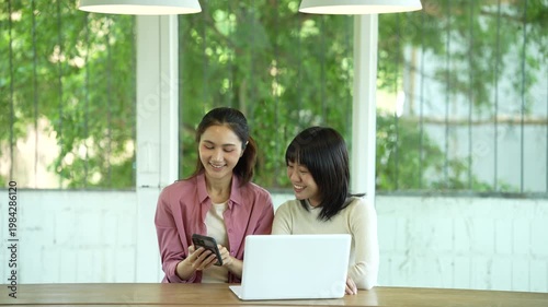 Two young women smiling and collaborating on a laptop in a bright modern workspace, representing teamwork, productivity, and a positive working atmosphere.