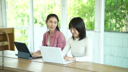 Two young women smiling and collaborating on a laptop in a bright modern workspace, representing teamwork, productivity, and a positive working atmosphere.