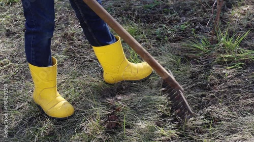 The video shows a woman in yellow rubber boots clearing a lawn with a rake. She collects old grass, fallen leaves, and dirt, working diligently to prepare the yard for spring growth.