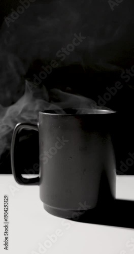 A matte black mug emits swirling white steam against a dark background, lit with dramatic contrast on a minimal surface, creating a moody, atmospheric scene with copy space
