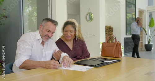 After agent call, Diverse couple sitting at table in modern home signing documents finalizing deal