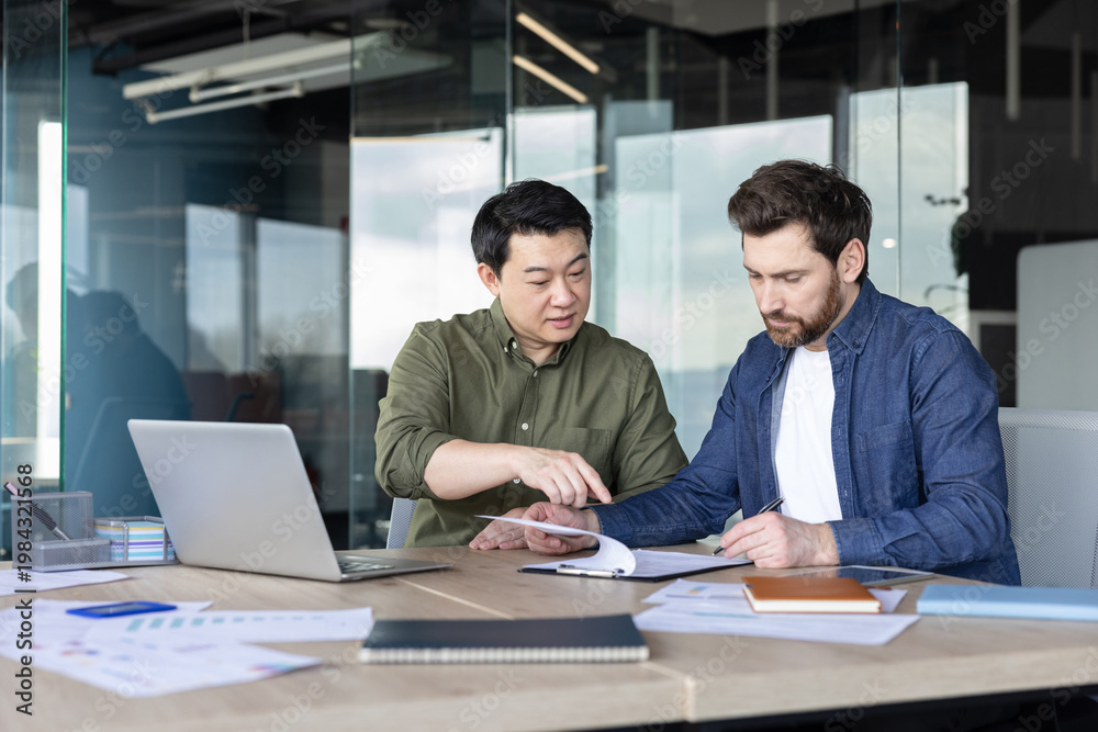 Obraz premium Two diverse businessmen are analyzing documents and discussing details around a desk in a modern office, working together on a strategic partnership or agreement